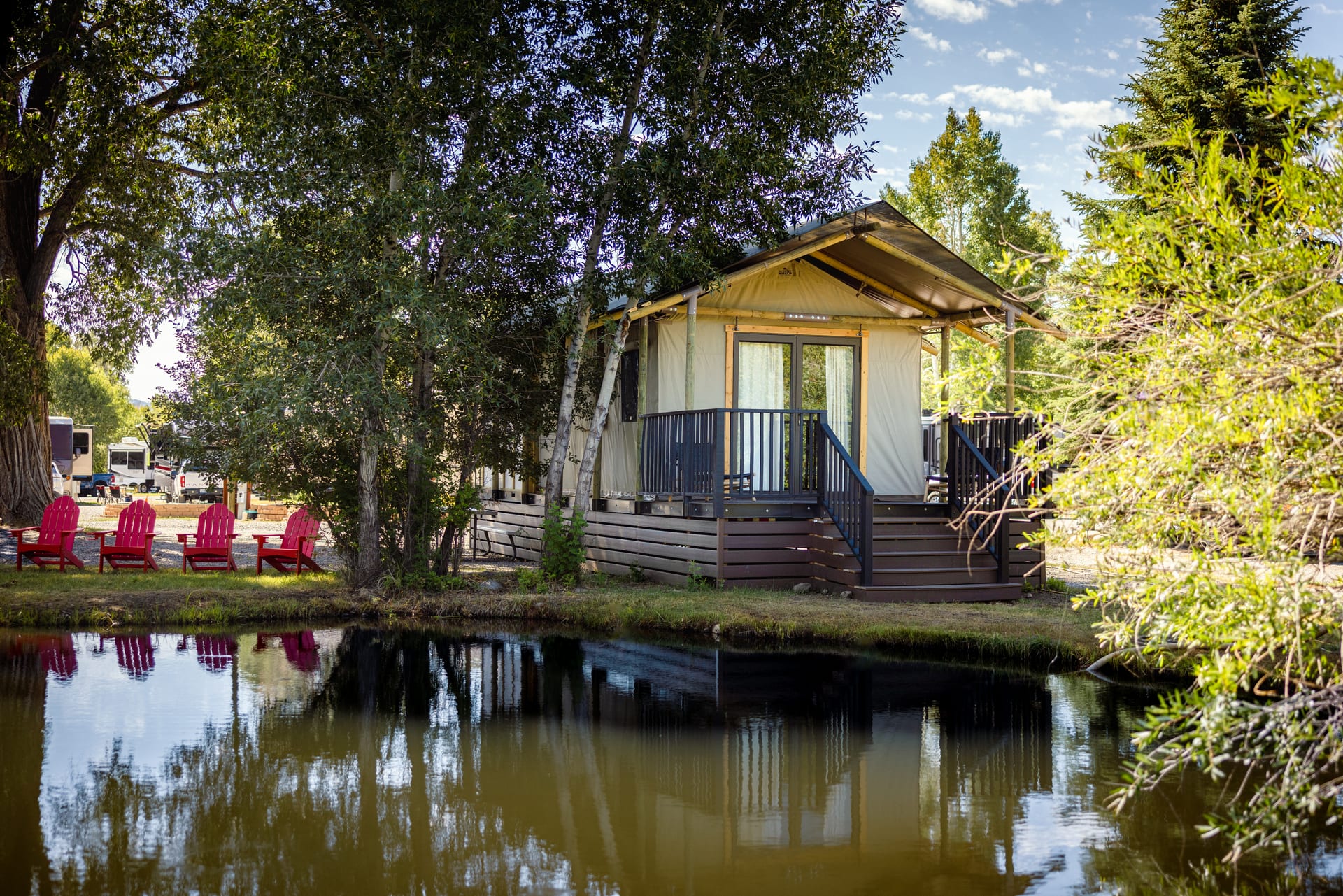 Glamping tents reflected in the pond with red adirondack chairs along the bank at Outdoorsy Bayfield